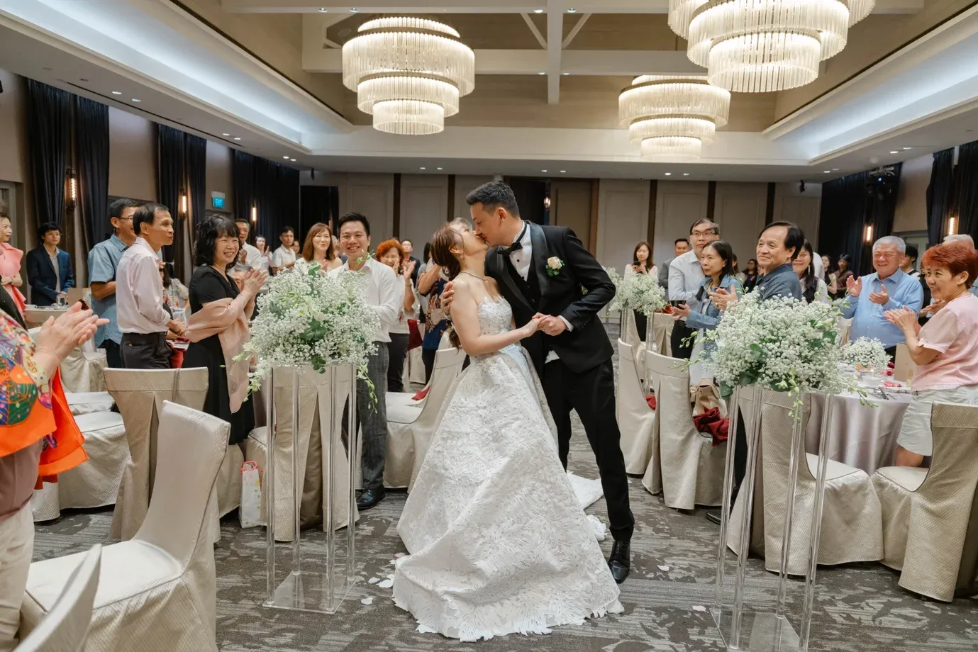 Couple kissing during their banquet march-in as guests applaud beneath the ballroom chandeliers