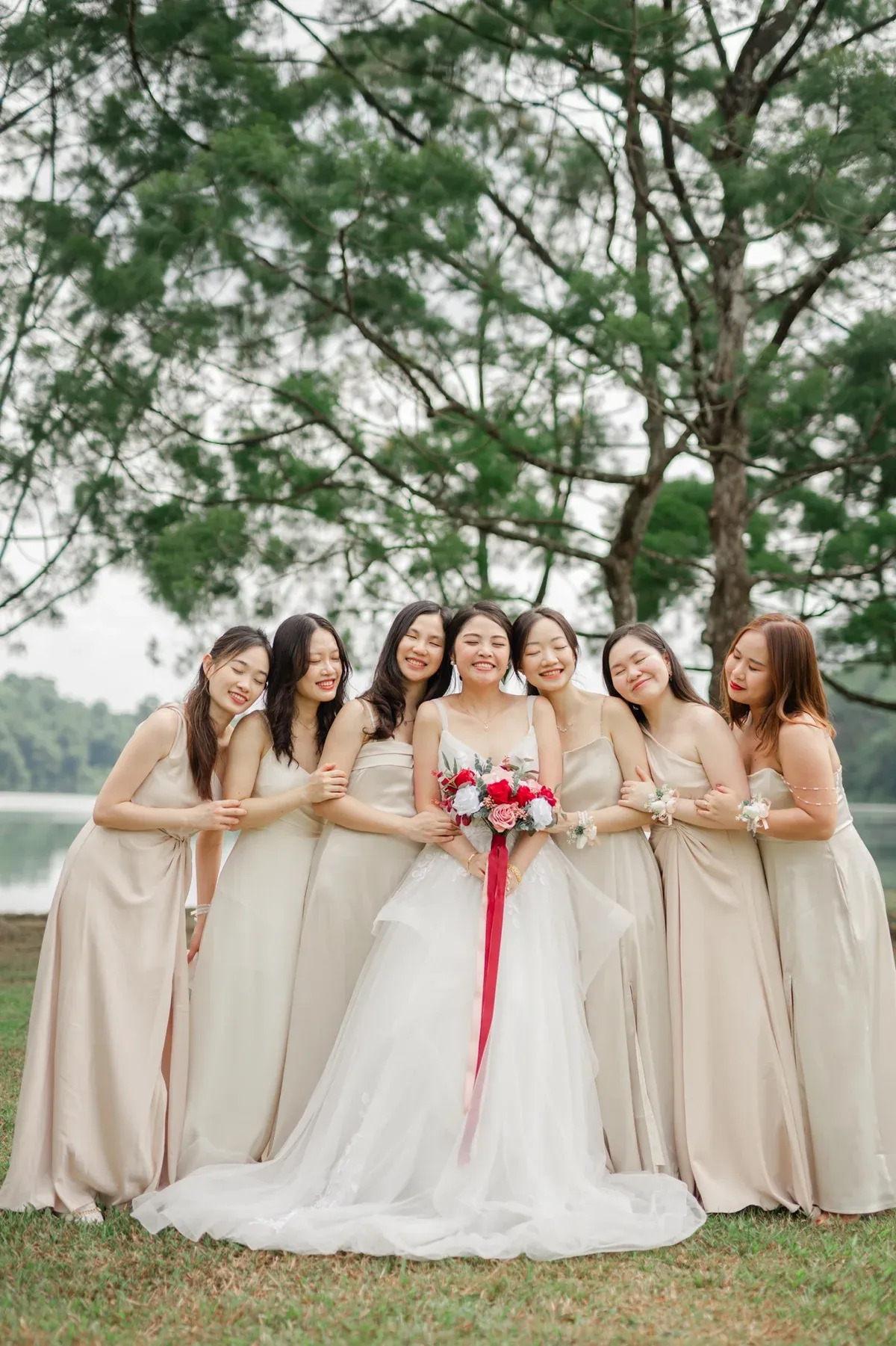 Bride laughing with her seven bridesmaids under casuarina trees beside a reservoir