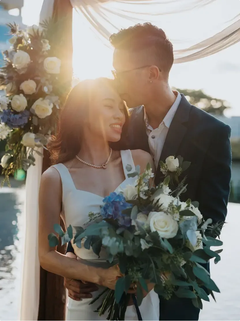 Backlit couple embracing under a floral arch at sunset, the bride holding her bouquet